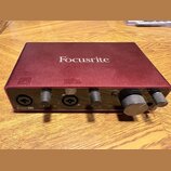 Červená audio rozhraní s nápisem Focusrite na horní straně. Na předním panelu jsou dva kombinované vstupy, dva ovládací knofíky zyska a velký knofík…