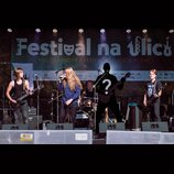 Fotografie zachycuje hudební kapelu na pódiu před černým bannerem s nápisem „Festival na ulici MULTIŽÁCROVÝ FESTIVAL V ULICÍCH PLZNĚ“. Vlevo stojí ky…