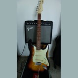 Elektrická kytara s trupem v barevnosti sunburst, bílým pickguardem a hnedým hmatníkem. Na hlavě kytary je vidět nápis Squier. Kytara stojí před kyta…
