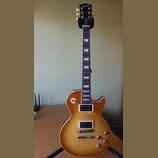 Elektrická kytara s trupem v žlutém gradientu (sunburst) a tmavým hmatníkem s trapezoidními vložkami. Na hlavě je bílý nápis Gibson. Nástroj má dva h…
