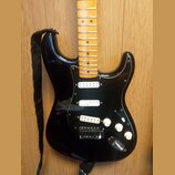 Černá elektrická kytara s tvarem typu Stratocaster a černým pickguardem. Má světle hnědý javorový hmatník a tři bílé snímače. Na pickguardu je vidite…