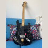 Černá elektrická kytara s mírně prohnutým tělem a světle hnědým hnedem hmatníkem. Na hlavě je nápis Squier Stratocaster. Tělo má černý pickguard, tři…