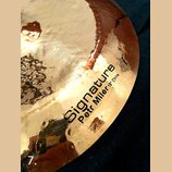 Detailní záběr na povrch mosazného činele s kovaným povrchem. Na čineli je černý nápis „Signature“ a pod ním menší text „Petr Milér 19" China“. Povrc…