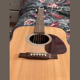 Akustická kytara s přírodním dřevem na desce a hnědým hmatníkem. Na desce je tmřečervený pickguard s mramorovým vzorem. Nástroj nemá žádné viditelné…