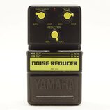 Černý kytarový pedál s nápisem NOISE REDUCER a označením modelu NR-100. Na horní části jsou tři žluté oválné ovladače s popisky THRESHOLD, DECAY a RE…