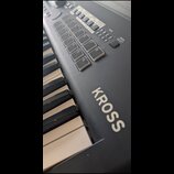 Detailní pohled na černý digitální klavír s bílými klávesami. Na korpusu je výrazný bílý nápis KROSS. Horní panel obsahuje sadu bílých čtvercových tl…