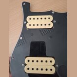 Černý plastový pickguard s dvěma humbuckery v krémové barvě. Snímače mají viditelné černé póly a jsou přišroubované k desce. Na povrchu pickguardu js…