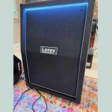 Černý reproduktorový box s černou textilní maskou a bílým rámečkem. Uprostřed masky je umístěn bílo-černý obdélníkový štítek s nápisem Laney. Povrch…
