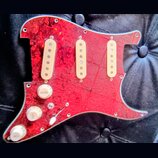 Červený pickguard s texturou připomínající mramor nebo plameny a stříbrným okrajem. Je na něm osazeno tři bílé snímače s viditelnými póly. V dolní čá…