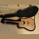 Akustická kytara s cutawayem v přirozené dřevěné barvě, umístěná v černém tvrdém kufru. Na hlavě hmatníku je viditelné logo „Takamine“. Povrch nástro…