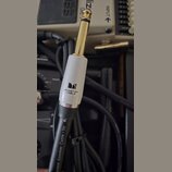 Fotografie zachycuje konektor typu jack v zlatém provedení s bílým plastovým krytem. Na krytu je černé logo ve tvaru písmene M a nápis MONSTER CABLE.…