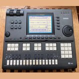 Šedý music sequencer značky YAMAHA, model QY700. Zařízení má v horní části displej s modrým podsvícením a pod ním řadu ovládacích tlačítek. V pravé č…