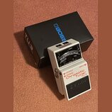 Bílý kytarový ladič s černým spátkem a ovládacími prvky. Na zařízení je uveden nápis BOSS, Chromatic Tuner TU-3 a OUTPUT BYPASS INPUT. Vedle ladiče j…