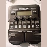 Černý multi-efektový procesor s nápisy G1 FOUR, ZOOM a Multi-Effects Processor. Zařízení má displej, čtyři otočné ovladače s popisky Lo, Mid, Hi a Vo…