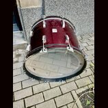 Basový buben v tmavě červené barvě s kovovým kováním. Povrch nástroje vykazuje viditelné opotřebení, škrábance a několik světlých skvrn. Na přední st…