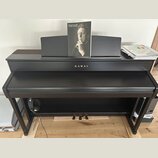 Černý digitální piano s uzavřenou klaviaturní víkem a integrovaným stojanem. Na přední straně je zlatý nápis KAWAI. Na notovém pultu je umístěn album…