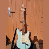 Světle modrá elektrická kytara s bílým pickguardem a javorovým hmatníkem. Na hlavě je viditelné logo Squier. Tělo kytary vykazuje opotřebení v podobě…