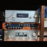 Tři stohované elektronické komponenty v regálu. Horní je stříbrný receiver s nápisy SONY, Digital Audio/Video Control Center a HDMI. Prostřední je or…
