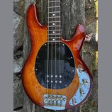 Elektrická basová kytara s hnědým slunečním odstínem (sunburst) a černým kruhovým pickguardem. Na horní části těla je vidět nápis Rickenbacker a chro…