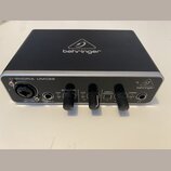 Černý audio rozhraní s hliníkovým stříbrným horním panelem. Na přední straně je nápis U-PHORIA UMC22 a logo behringer, stejně jako na horní části zař…