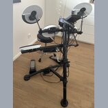 Elektronická bicí souprava na černém kovovém stojanu. Na činelech je vidět nápis Roland, na sluchátkách nápis audio-technica. V pozadí je černý buben…