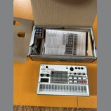 Na fotografii je bílý sampler KORG volca sample v originální krabici. Za přístrojem je otevřený kartonový obal, ve kterém se nachází uživatelský manu…