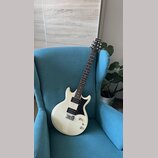 Bílá elektrická kytara s černým pickguardem a hnedým hmatníkem. Na hlavě je viditelný nápis Brandia. Nástroj má dva snímače typu humbucker a jeden je…