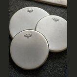 Tři bílé kruhové bubnové blány s kovovými obručemi. Každá blána má v horní části černé logo REMO a pod ním nápis EMPEROR® WHITE COATED MADE IN U.S.A.…