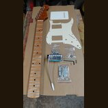 Sada komponentů pro elektrickou kytaru položená na hnědé kartonové ploše. Obsahuje dřevěný krk s hnědým lakem, bílý plastový pickguard a bílou krytku…