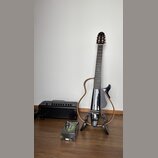 Na fotografii je černá elektrická kytara s atypickým dutým korpusem v podobě obrysu, umístěná na černém stojanu. Vedle kytary stojí malý černý komboz…