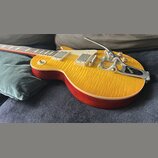Elektrická kytara s žlutým plamenovitým topem a červenými boky. Na kytaru je instalován chromovaný Bigsby vibrační systém s nápisem „Bigsby“. Nástroj…
