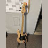 Elektrická basová kytara s přírodním dřevěným povrchem těla a hmatníku. Nástroj má černý pickguard, dva snímače a čtyři ladicí kolíky na hlavě. Na hl…