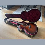 Akustická kytara s cutawayem v tmavě hnědém sunburst provedení, umístěná v otevřeném černém tvrdém pouzdře s červeným plyšovým vnitřkem. Na hlavě hma…