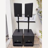 Dva černé reproduktory umístěné na černých stojaních, které jsou zapuštěny do dvou černých subwooferů. Všechny čtyři komponenty mají na přední straně…