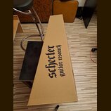 Kartonový obal v hnědé barvě pro hudební nástroj. Na povrchu je černý nápis Schecter guitar research. Obal vykazuje mírné pomačkání v dolní části.