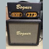 Kytarový zesilovač a reproduktorové boxy značky Bogner. Horní jednotka má zlatý ovládací panel s nápisy BARCELONA, volume, gain, bass a treble. Předn…
