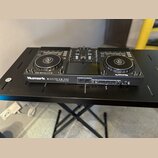 Černý DJ 컨트롤러 Numark MIXSTREAM PRO umístěný na černém stole. Na přední straně je vidět bílý nápis Numark MIXSTREAM PRO a menší text pod ním, který je…