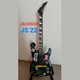 Černá elektrická kytara s hrotovitou hlavou, na které je bílé logo Jackson. Tělo kytary je polepeno různými nálepkami, včetně log Monster Energy a Ho…