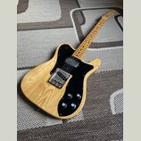Elektrická kytara s přírodním dřevem a černým pickguardem. Na hlavě hnedku je viditelné logo Fender. Kytara má dva humbuckery, tři ovládací knofíky a…