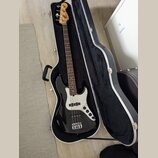 Černá elektrická basová kytara v otevřeném tvrdém kufru s černým vnitřkem. Na hlavě nástroje je viditelné logo Fender. Kytara má bílý pickguard, čtyř…