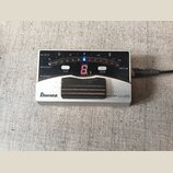 Stříbrný digitální ladič s černým ovládacím panelem a gumovým pedálem. Na panelu jsou nápisy DC IN, OUTPUT, DRYOUT, INPUT a tlačítka FLAT a CALIB. V…