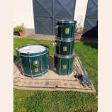 Sada bubnů v tmavě zelené barvě s kovovým hardwarem. Na každém z bubnů je žlutý čtvercový štítek s nápisem „FIRE SHELL“ a symbolem radioactivity. Sad…