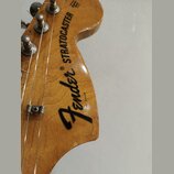 Detail hlavy kytary z dřeva s lakovaným povrchem. Na hlavě je černý nápis Fender® STRATOCASTER a malé vertikální sériové číslo 3143028. Jsou vidět ko…