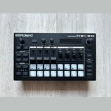 Černá groovebox s nápisy Roland a GROOVEBOX MC-101. Zařízení obsahuje displej, několik otočných ovladačů a bílá tlačítka s různými označením, včetně…