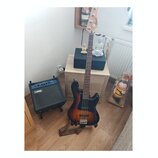 Elektrická basová kytara s tmavě hnědým sunburst povrchem a hmatníkem z tmavého dřeva. Na hlavě nástroje je bílý nápis Cort. Vedle kytary stojí malý…