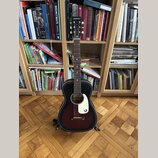 Akustická kytara s tmavě červeným gradientem (sunburst) a bílým plektrem. Na hlavě hmatníku je bílý nápis GRETSCH a na plektru bílé logo G. Nástroj j…