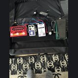 V černé přepravní tašce je umístěno několik efektových pedálů propojených kabely. Zleva jsou vidět červený pedal s nápisem "Electro-Harmonix" a "Sava…