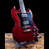 Elektrická kytara s červeným lakem a černým pickguardem, umístěná na černém stojanu. Nástroj má dva humbuckery a čtyři ovládací knoflíky. Povrch laku…