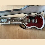 V otevřeném černém kufru s bílou vnitřní výplní leží červená elektrická kytara. Na hlavě nástroje je viditelné logo Gibson. Kytara má dva humbuckery,…