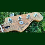 Záběr na hlavu basové kytary z přírodního dřeva s viditnou masivní strukturou. Na zadní straně je černý nápis Fender® JAZZ BASS®. Jsou vidět čtyři ko…